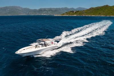 A white speedboat cuts through the deep blue ocean, leaving a frothy wake, with a lush green coastline and distant mountains under a clear sky in the background.