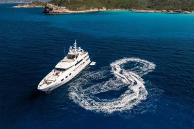 A large white yacht sails through deep blue water, leaving a foamy figure-eight wake, with a lush green coastline visible in the distance.