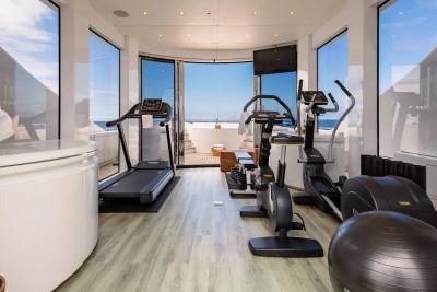 A well-equipped gym with treadmills and exercise bikes is situated on a yacht, with a stunning ocean view visible through large windows in the background.