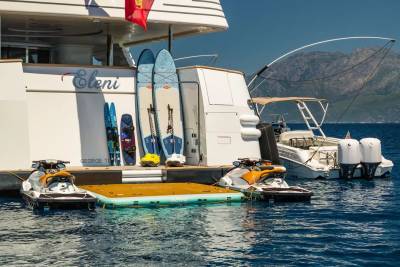 Two jet skis are docked at a floating platform in front of a large yacht, with a smaller boat and mountainous coastline in the background.
