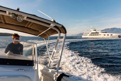 A person steers a small boat through choppy water, leaving a white wake, with a large white yacht sailing in the distance against a clear blue sky and distant hills.