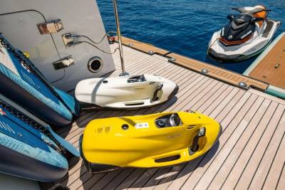 Two colorful underwater scooters and inflatable paddleboards rest on a yacht's deck, with a jet ski and the blue ocean visible in the background.