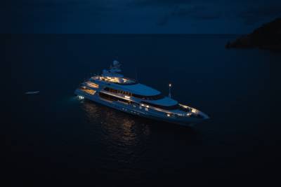 A luxurious yacht is illuminated at night in calm, dark water, with a dimly lit coastline visible in the distance.
