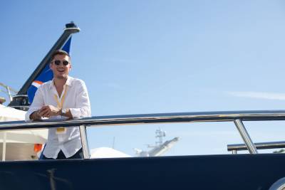 A man in a white shirt smiles on a yacht's deck with a clear blue sky and another boat in the background.