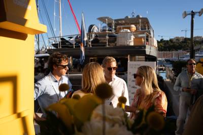 In the foreground, a group of people are gathered near a yellow structure, while in the background, a large yacht is docked in a harbor with buildings on a hillside.