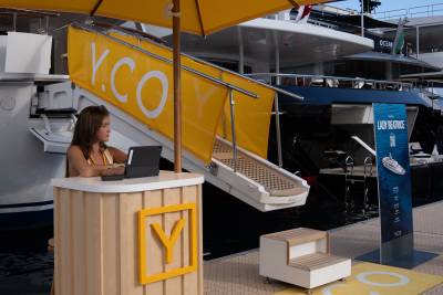 A woman works at a Y.CO. branded stand in the foreground, with a large yacht named "LADY BEATRICE" docked behind her.