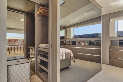 A modern closet with a woven hamper in the foreground opens to a bedroom with a large mirrored wall reflecting a bed and a window with a view of the sky.