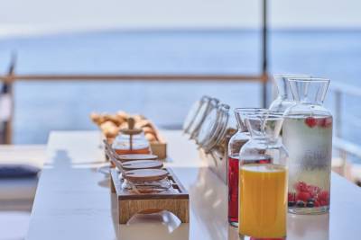 A breakfast spread of juices, honey, and granola sits on a table with a blurred ocean and sky in the background.