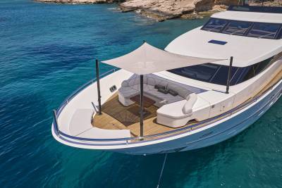A luxurious white yacht with a shaded seating area in the foreground floats on clear turquoise water near a rocky, tree-lined coastline in the background.