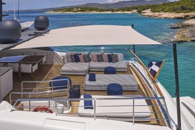 A luxurious yacht deck with comfortable seating and a dining area is anchored in a clear turquoise bay with a hilly, tree-lined coastline in the background.