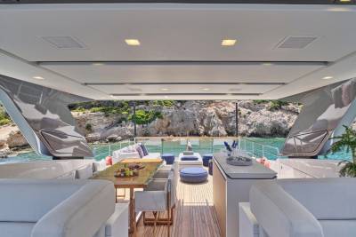 A luxurious yacht's open-air deck with a dining area in the foreground and a rocky coastline with clear blue water in the background.