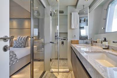 A modern yacht bathroom with a double vanity in the foreground and a glimpse of a bedroom through a glass door in the background.