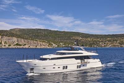A sleek white yacht cruises on calm blue water with a rocky, tree-covered hillside under a bright blue sky in the background.