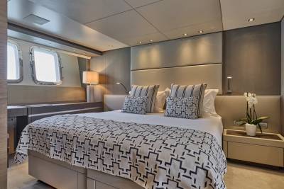 A luxurious yacht bedroom features a king-sized bed with patterned bedding in the foreground and porthole windows with a desk in the background.