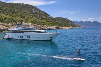A person on a paddleboard glides across clear blue water in the foreground, with a large white yacht anchored near a lush, green, mountainous coastline in the background.