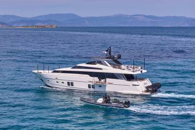 A sleek white yacht cruises on calm blue water with a small inflatable boat in the foreground, while a hazy, mountainous coastline stretches across the background.