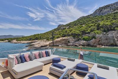 A luxurious yacht deck with plush seating in the foreground overlooks a serene bay with a lush, green mountainside in the background.