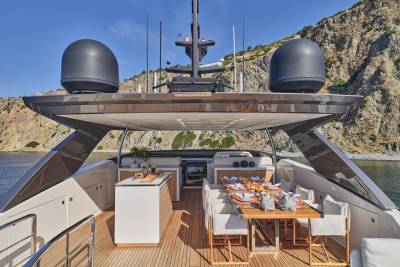 A luxurious yacht deck with a dining area and outdoor kitchen is anchored near a rocky, sun-drenched coastline.