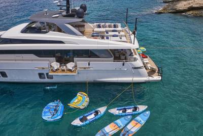 A luxurious white yacht floats in clear turquoise water, surrounded by inflatable water toys and paddleboards, with a rocky coastline visible in the background.