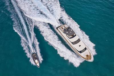 A large yacht and a smaller boat speed across turquoise water, leaving white wakes behind them.