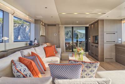 A luxurious yacht interior with a white sectional sofa adorned with orange and blue patterned pillows in the foreground, and a scenic ocean view with rocky cliffs visible through large windows in the background.