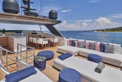 A luxurious yacht deck with white lounge chairs and blue cushions in the foreground is set against a backdrop of the sparkling blue ocean and a distant, tree-covered coastline under a clear sky.