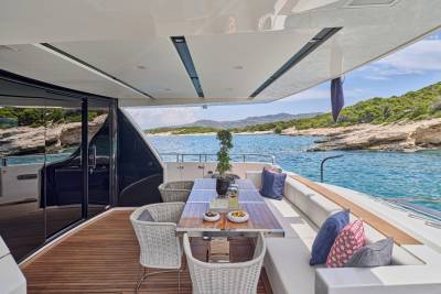 A luxurious yacht deck with a dining table and seating in the foreground overlooks a picturesque coastline with calm blue waters in the background.