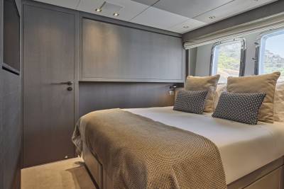 A neatly made bed with plush pillows and a textured throw occupies the foreground, while large windows in the background reveal a glimpse of a sunny, green landscape.