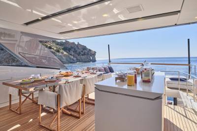 A beautifully set dining table and bar area on a yacht's deck overlooks a rocky coastline and calm blue sea.