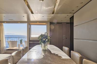 A marble dining table with a floral centerpiece sits in the foreground of a luxurious yacht interior, with a bright ocean view visible through large windows in the background.