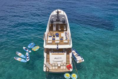 A large white yacht floats in clear turquoise water, surrounded by paddleboards and inflatable tubes, with a small dinghy tied to its side.