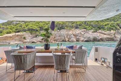 Wicker chairs surround a wooden table on a yacht's deck, with a lush, rocky coastline visible in the background.