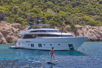 A woman on a paddleboard glides on clear blue water in front of a large white yacht anchored near a lush, green, rocky coastline.