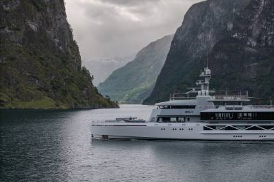 A sleek, modern yacht sails on calm water between towering, verdant cliffs under a cloudy sky.