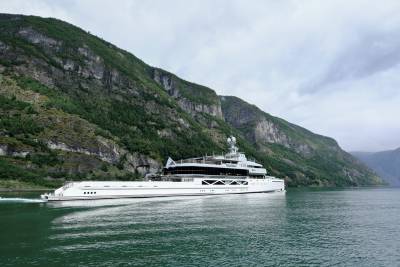 A large white yacht cruises on calm water in the foreground, with steep, green, tree-covered mountains rising dramatically in the background.