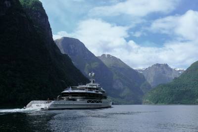 A large white yacht cruises through a calm body of water, dwarfed by the majestic, green-covered mountains that rise steeply on either side.