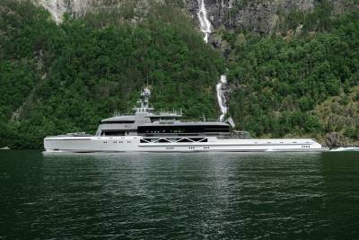 A sleek, modern yacht cruises on calm water in the foreground, with a lush, green mountainside featuring a cascading waterfall in the background.