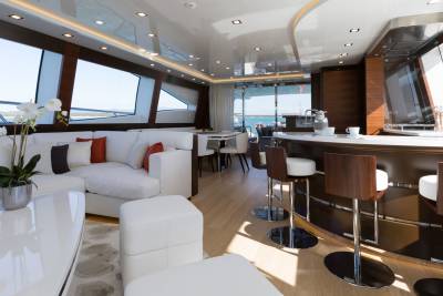A luxurious yacht interior features a white sectional sofa and ottomans in the foreground, with a polished bar and dining area visible in the background through large windows.