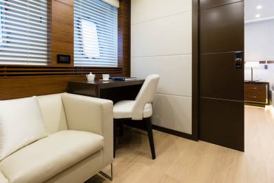 A cream-colored sofa sits in the foreground next to a desk with a chair, while a bedroom with a lit bedside lamp is visible in the background through an open doorway.