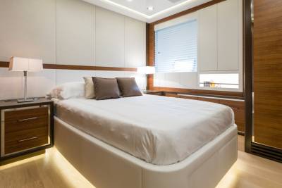 A luxurious bedroom with a white bed and bedside table in the foreground, and a window with blinds and wooden cabinets in the background.