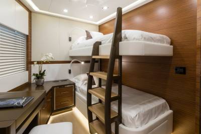 A bunk bed with white linens is in the foreground, while a wooden-paneled wall and a desk with blinds are in the background.