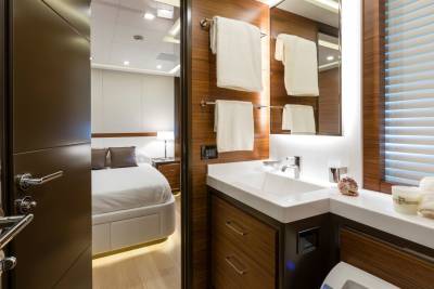 A modern yacht bathroom with a white sink and wooden cabinets is in the foreground, while a neatly made bed in a bedroom is visible in the background through an open doorway.