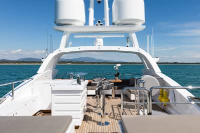 A luxurious yacht's deck with comfortable seating and tables is visible in the foreground, while the calm blue ocean stretches to a distant, tree-lined shore under a clear sky in the background.