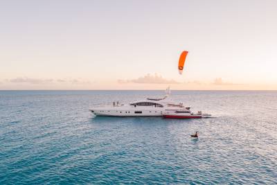 A large white yacht with a red hull is anchored in calm turquoise waters, while a person kitesurfs in the foreground against a soft, hazy sky.