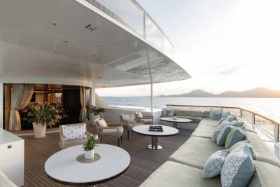 A spacious, sun-drenched yacht deck with comfortable seating and tables in the foreground overlooks a calm sea and distant islands bathed in the warm glow of sunset.