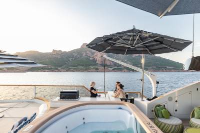 A luxurious yacht's deck features a steaming hot tub in the foreground and a scenic coastal mountain range in the background.