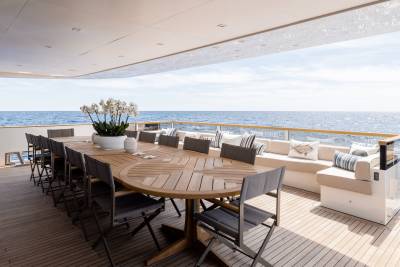 A large oval wooden dining table with chairs is set on a yacht deck, with the ocean and sky visible in the background.