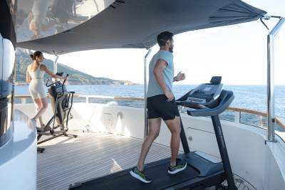 A man runs on a treadmill and a woman uses an elliptical machine on a yacht's deck, with the ocean and coastline visible in the background.