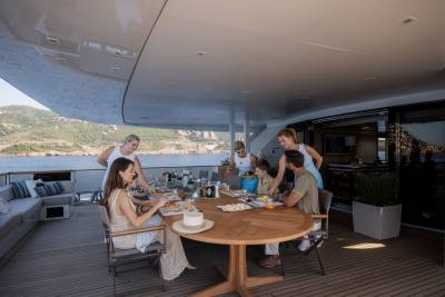 A family enjoys a meal on a yacht's deck with a scenic coastal landscape in the background.