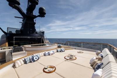 A spacious, sun-drenched deck with plush seating and small tables is in the foreground, while a large, dark radar mast and the vast blue ocean stretch into the background under a clear sky.